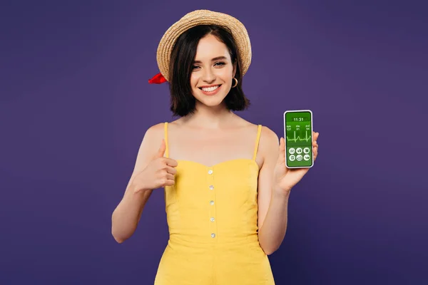 Sonriente chica bonita en sombrero de paja que muestra el pulgar hacia arriba mientras sostiene el teléfono inteligente con aplicación sanitaria aislada en púrpura - foto de stock