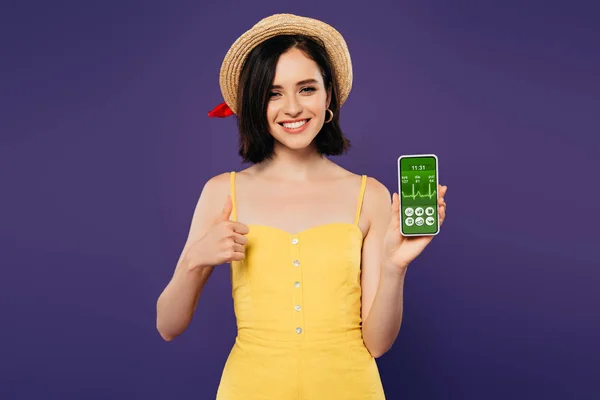 Sourire jolie fille en paille chapeau montrant pouce vers le haut tout en tenant smartphone avec application de soins de santé isolé sur violet — Photo de stock