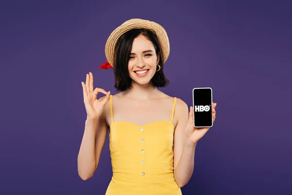 KYIV, UCRANIA - 3 DE JULIO DE 2019: niña bonita y sonriente con sombrero de paja sosteniendo el teléfono inteligente con la aplicación HBO y mostrando un signo aceptable aislado en púrpura - foto de stock