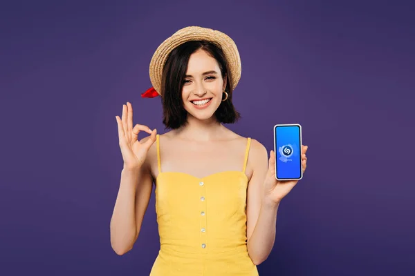 KYIV, UCRAINA - LUGLIO 3, 2019: bella ragazza sorridente in cappello di paglia che tiene smartphone con app Shazam e mostra un segno ok isolato sul viola — Foto stock