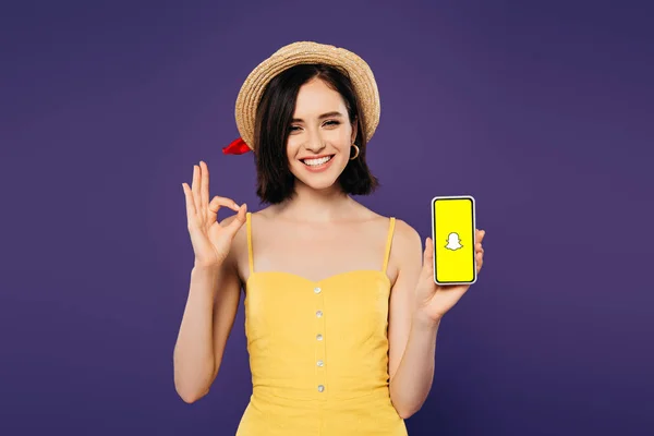 KYIV, UKRAINE - 3 juillet 2019 : jolie fille souriante en chapeau de paille tenant smartphone avec application Snapchat et montrant ok signe isolé sur violet — Photo de stock