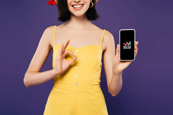 KYIV, UKRAINE - JULY 3, 2019: cropped view of smiling girl in holding smartphone with deezer logo and showing ok sign isolated on purple — Stock Photo