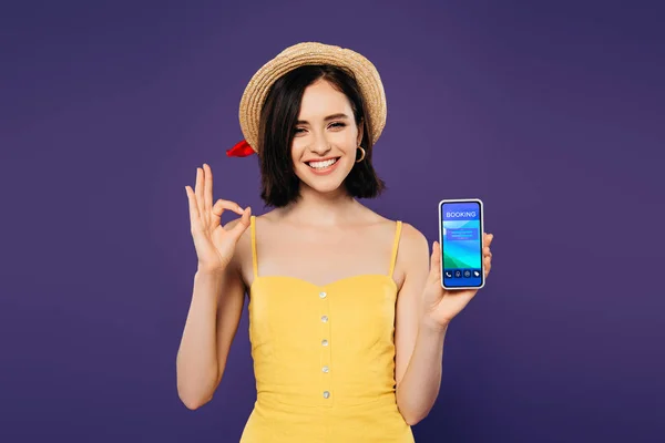 Sorridente bella ragazza in cappello di paglia in possesso di smartphone con app di prenotazione e mostrando segno ok isolato sul viola — Foto stock