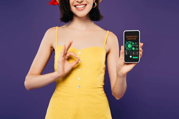 Cropped view of smiling girl in holding smartphone with marketing analysis app and showing ok sign isolated on purple — Stock Photo