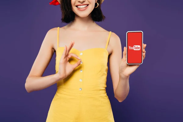 Kyiv, Ukraine - 3. Juli 2019: Ausgeschnittene Ansicht eines lächelnden Mädchens, das ein Smartphone mit Youtube-Logo in der Hand hält und ein Ok-Zeichen auf Lila zeigt — Stockfoto