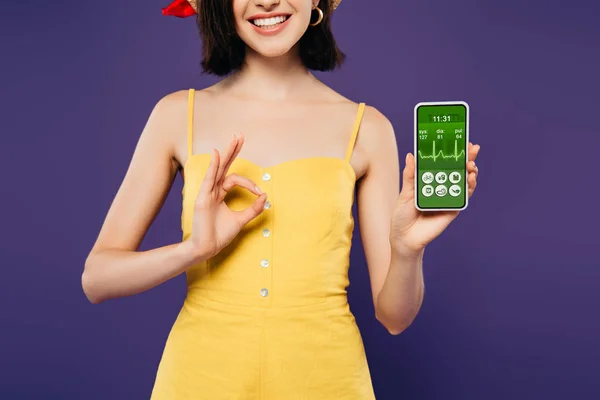 Обрізаний вид усміхненої дівчини, що тримає смартфон із додатком охорони здоров'я та показує знак ОК ізольовано на фіолетовому — стокове фото
