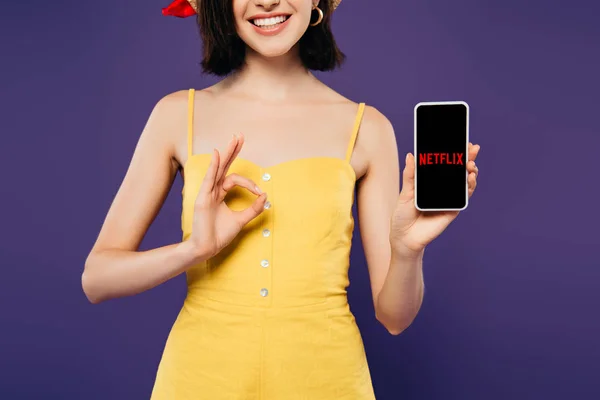 КИЕВ, УКРАИНА - 3 ИЮЛЯ 2019 года: обрезанный вид улыбающейся девушки в держании смартфона с логотипом netflix и показ ok знак изолирован на фиолетовый — стоковое фото