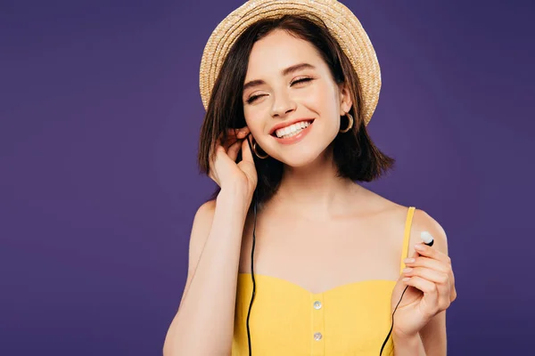 Sorridente bella ragazza in cappello di paglia con gli occhi chiusi mettere gli auricolari isolati su viola — Foto stock