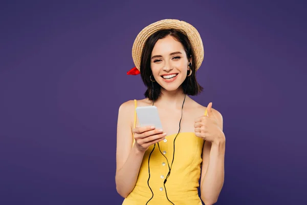 Smiling pretty girl in straw hat listening music in earphones on smartphone and showing thumb up isolated on purple — Stock Photo