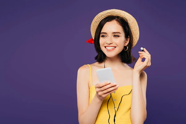 Sonriente chica bonita en sombrero de paja escuchando música en los auriculares en el teléfono inteligente y mirando hacia otro lado aislado en púrpura - foto de stock