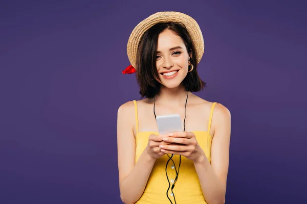 Sonriente chica bonita en sombrero de paja escuchar música en los auriculares en el teléfono inteligente aislado en púrpura - foto de stock