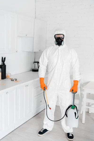 Exterminator in protective mask and uniform holding toxic equipment while standing near kitchen cabinet — Stock Photo