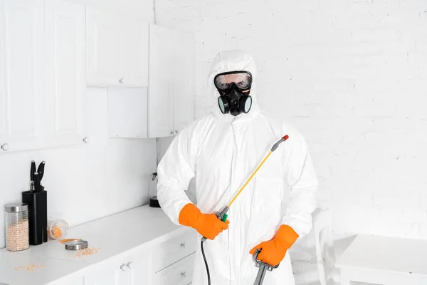 Exterminator in protective mask and uniform holding toxic equipment near kitchen cabinet — Stock Photo