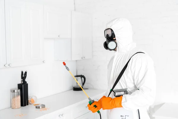 Exterminator in protective mask holding toxic equipment near kitchen cabinet — Stock Photo