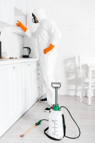 Exterminador en máscara protectora de pie con la mano en la cadera cerca del gabinete de cocina - foto de stock