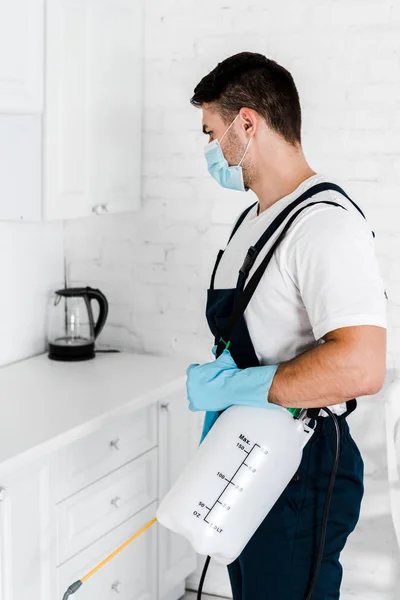 Exterminador em máscara protetora e uniforme segurando spray tóxico em pé na cozinha — Fotografia de Stock