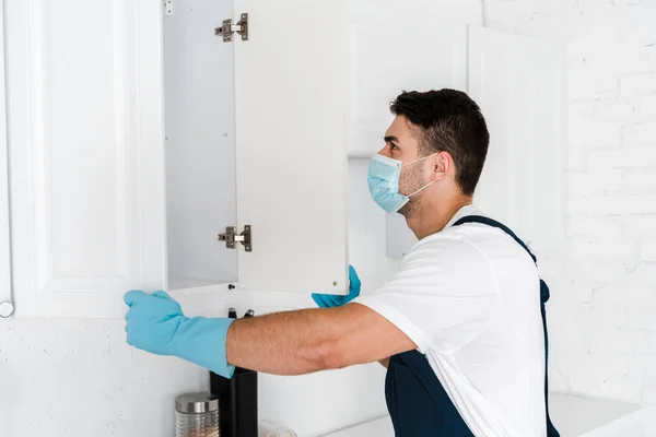 Exterminator in uniform looking at kitchen cabinet — Stock Photo