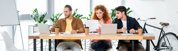 Панорамный снимок трех друзей, использующих ноутбук и делающих бумажную работу в офисе — стоковое фото