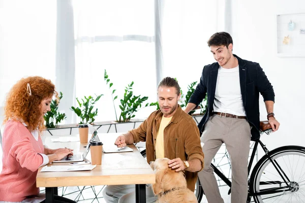 Mulher atraente usando laptop e amigos acariciando golden retriever no escritório — Fotografia de Stock