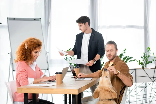 Friends using laptop, doing paperwork and feeding golden retriever — Stock Photo