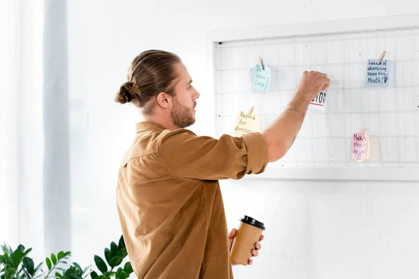 Gutaussehender Mann im Hemd, der an der weißen Tafel eine Karte aufsetzt und Pappbecher im Büro hält — Stockfoto