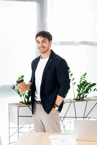 Bonito e sorridente homem segurando copo de papel e olhando para a câmera no escritório — Fotografia de Stock