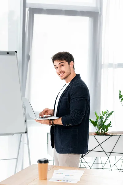 Hombre guapo y sonriente en camisa sosteniendo portátil y mirando a la cámara en la oficina - foto de stock