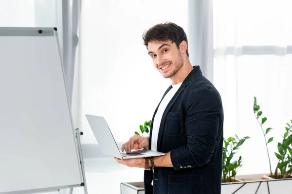 Hombre guapo y sonriente en camisa sosteniendo portátil y mirando a la cámara en la oficina - foto de stock