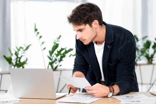 Hombre guapo haciendo papeleo y mirando el ordenador portátil en la oficina - foto de stock