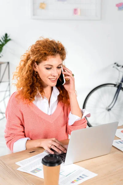 Atractiva mujer de negocios en suéter rosa hablando en el teléfono inteligente y utilizando el ordenador portátil en la oficina - foto de stock