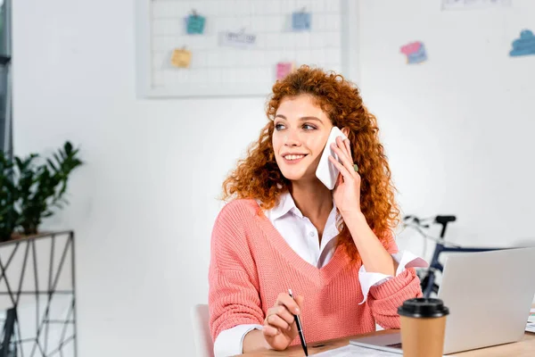 Attractive and smiling businesswoman in pink sweater talking on smartphone in office — Stock Photo
