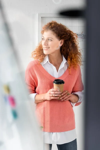 Messa a fuoco selettiva di attraente e sorridente donna d'affari in maglione rosa in possesso di tazza di carta — Foto stock