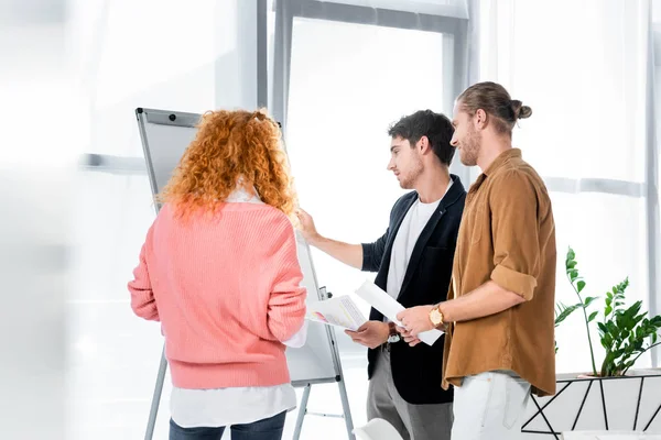 Drei Freunde, die im Büro Papierkram erledigen und Flipchart ansehen — Stockfoto