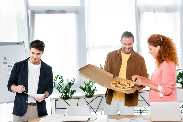 Улыбающиеся друзья берут пиццу из коробки и делают бумажную работу — стоковое фото