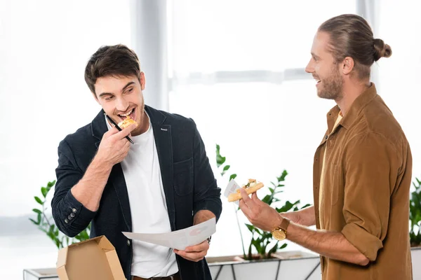 Two smiling friends eating pizza and holding paper in office — Stock Photo