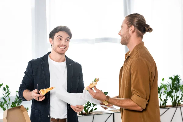 Two smiling friends holding pizza slices and papers in office — Stock Photo