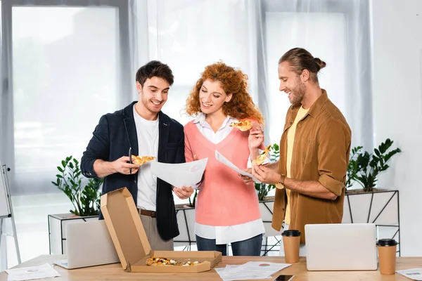 Tres amigos sonrientes sosteniendo rebanadas de pizza y haciendo papeleo en la oficina - foto de stock