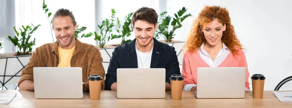 Plano panorámico de tres amigos sonrientes usando computadoras portátiles en la oficina - foto de stock