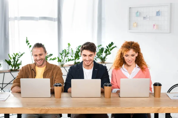 Tres amigos sonrientes sentados en la mesa y usando computadoras portátiles en la oficina - foto de stock
