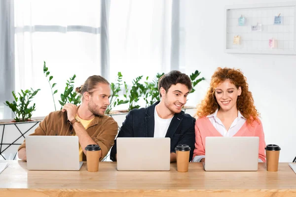 Tres amigos sonrientes sentados en la mesa y usando computadoras portátiles en la oficina - foto de stock