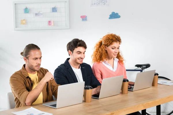 Three smiling friends sitting at table and using laptops in office — Stock Photo