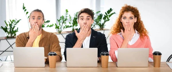 Panoramaaufnahme von drei schockierten Freunden, die am Tisch sitzen und Gesichter verdecken — Stockfoto