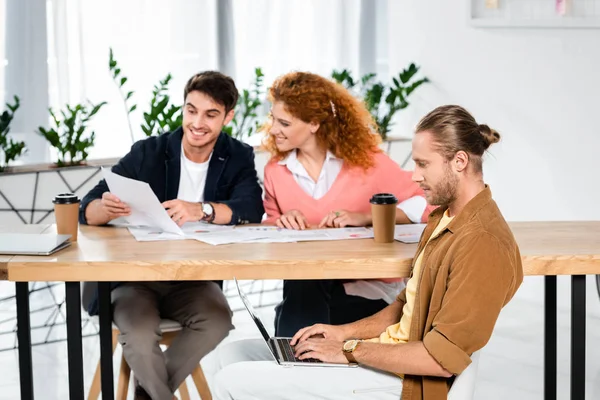 Tres amigos sonrientes haciendo papeleo y utilizando el ordenador portátil en la oficina - foto de stock