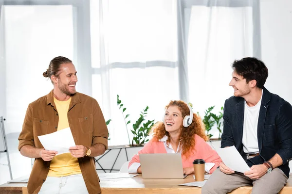 Tres amigos sonrientes haciendo papeleo y mirándose en la oficina - foto de stock