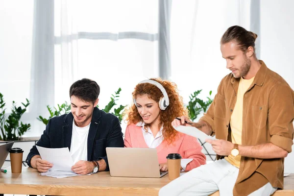 Tres amigos sonrientes haciendo papeleo y utilizando el ordenador portátil en la oficina - foto de stock