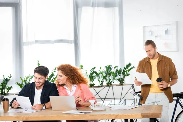 Tres amigos sonrientes haciendo papeleo y sentados en la mesa en la oficina - foto de stock