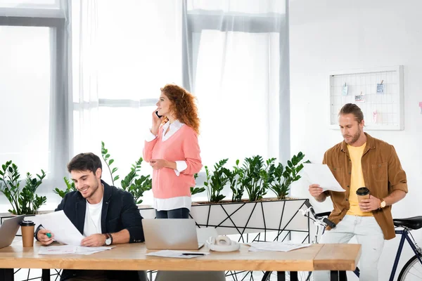 Tres amigos haciendo papeleo y hablando en smartphone en la oficina - foto de stock