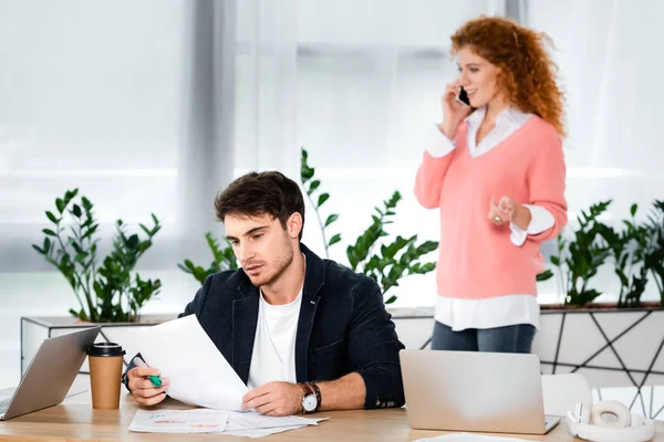 Избирательный фокус мужчины, делающего бумажную работу, и женщины, говорящей на смартфоне в офисе — стоковое фото