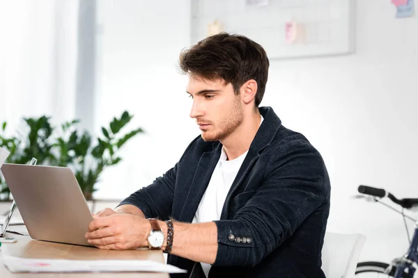Hombre de negocios guapo en camisa usando el ordenador portátil y sentado en la mesa en la oficina - foto de stock