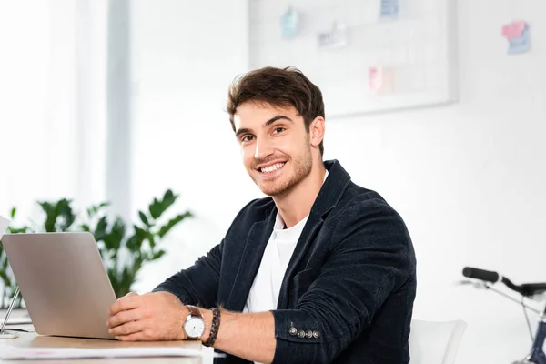 Красивий і усміхнений бізнесмен в сорочці, використовуючи ноутбук і дивлячись на камеру в офісі — стокове фото